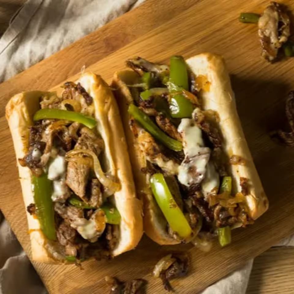 Two Philly cheesesteak sandwiches with steak, onions, peppers, and melted cheese on a wooden board.