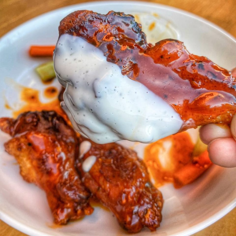A hand dips a saucy Milans Pizza & Wings chicken wing into a creamy white sauce.