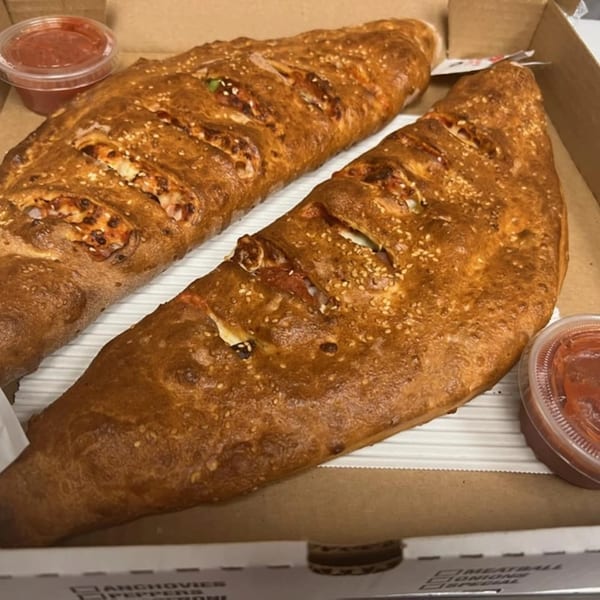 Two golden-brown calzones with sesame seeds and two cups of marinara sauce in a pizza box.