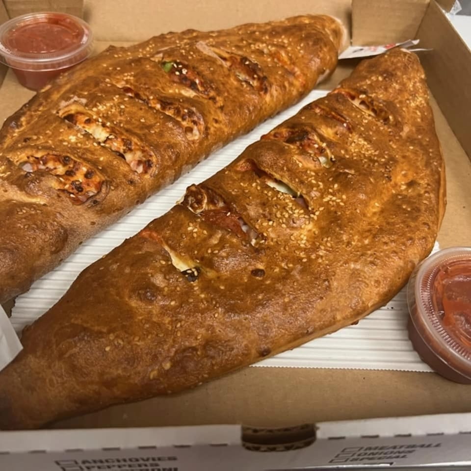 Two golden-brown calzones with sesame seeds and two cups of marinara sauce in a pizza box.